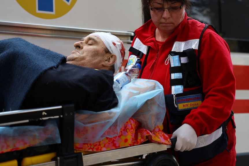 Next to an ambulance vehicle, a medic pulls a stretcher on which lies a man with bloodied bandages around his head.