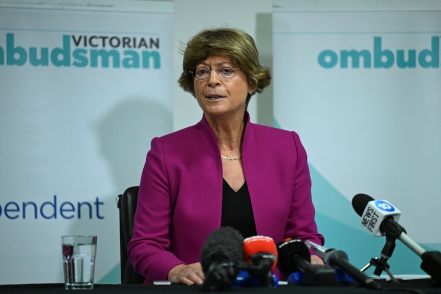 Deborah Glass sitting at a table in front of TV station microphones with a Victorian Ombudsman sign behind her.