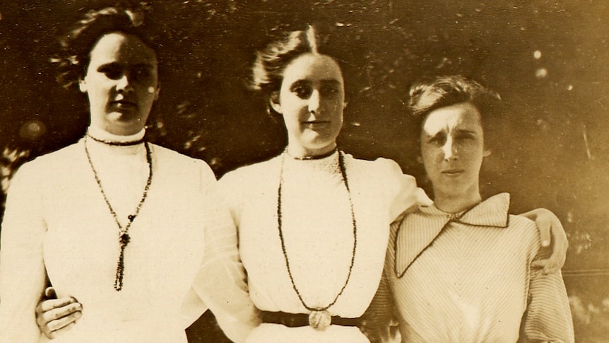 An historic photo of three women with their arms around each other, all in white long dressed. Vera Deakin stands in the middle.
