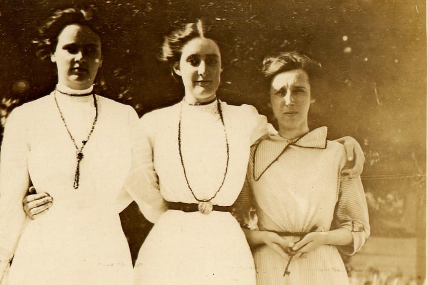An historic photo of three women with their arms around each other, all in white long dressed. Vera Deakin stands in the middle.