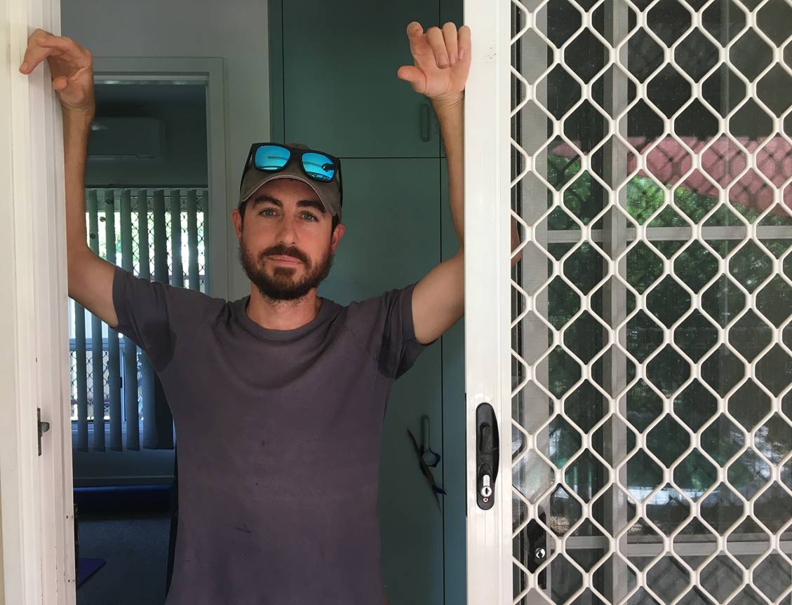 A man wearing a hat and sunglasses leans against a screen door.