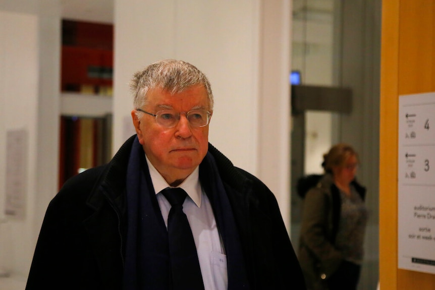 Didier Lombard walks down a corridor wearing a dark suit and glasses.