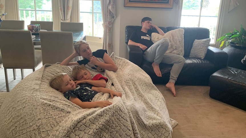 Three young children lie under a blanket on a couch and an older child sits on a separate chair in a family home