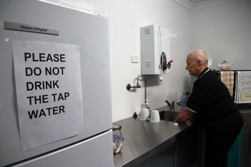 A woman stands at a sink washing dishes near a fridge with a sign warning people not to drink the tap water.