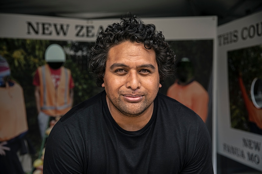 A man with dark curly hair smiles while standing in front of large boards with photographs and text.