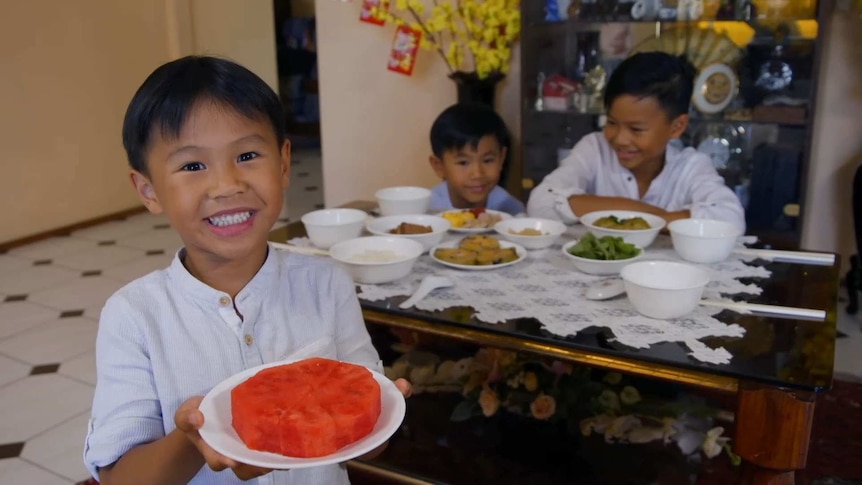 Boy holding up a plate with watermelon on it with his brothers sitting at a table behind him
