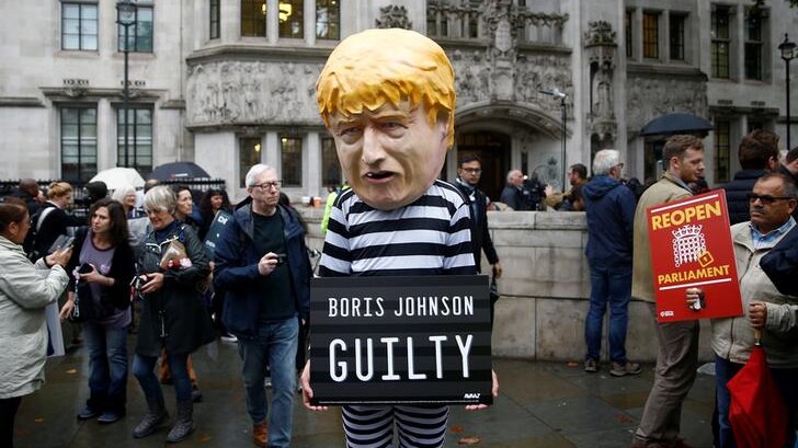 A man in a black and white prison outfit with a Boris Johnson bobble head mask on holds a sign reading: "Boris Johnson guilty".