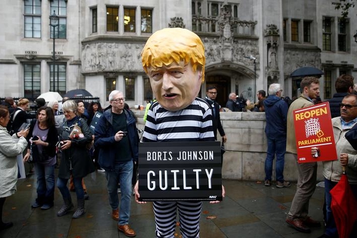 A man in a black and white prison outfit with a Boris Johnson bobble head mask on holds a sign reading: "Boris Johnson guilty".