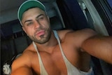 A man wearing a hat and singlet sits in a car taking a selfie on his phone