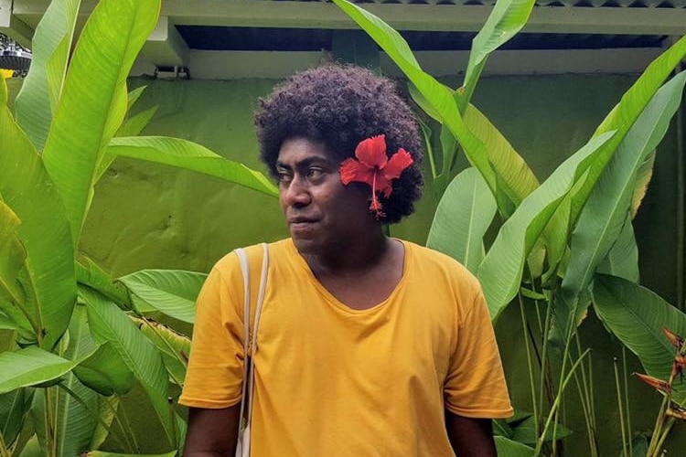 A person with short dark afro hair wearing a yellow top standing beside green banana leaves 