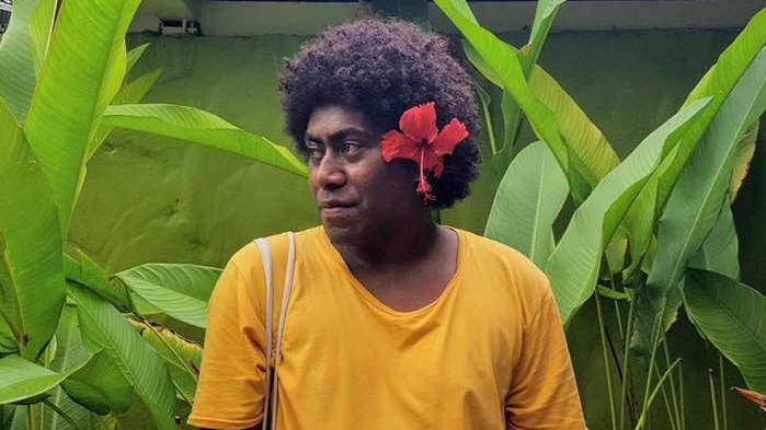 A person with short dark afro hair wearing a yellow top standing beside green banana leaves 