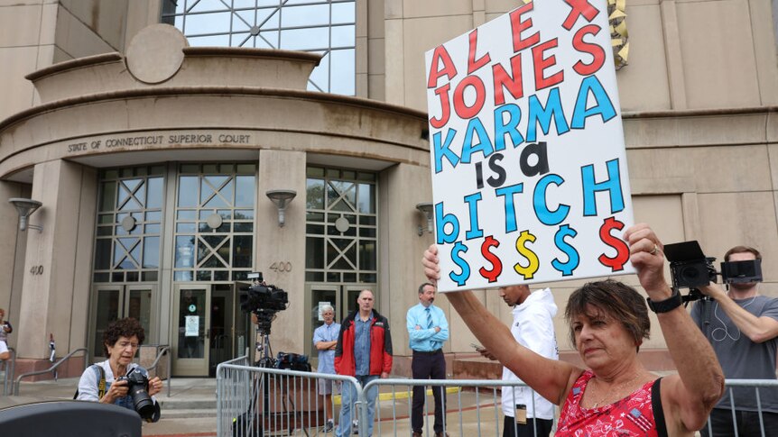 A woman outside courthouse holding sign "Alex Jones Karma is a bitch"