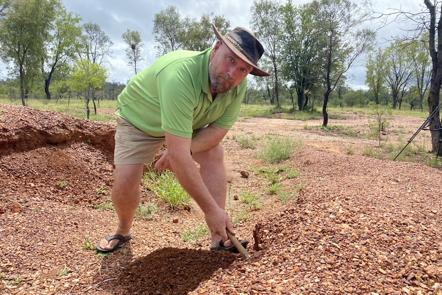 A man with a green shirt digs with a hammer for gems. He is wearing a hat.