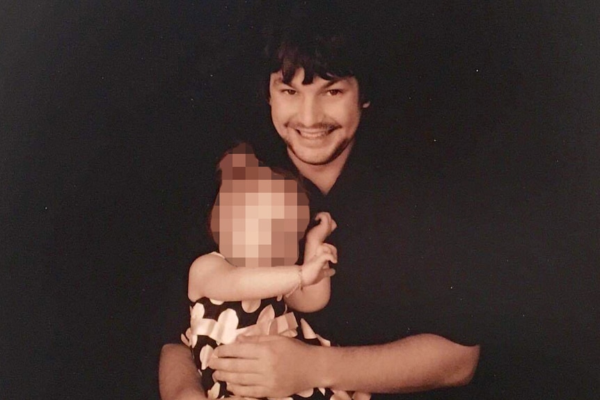 Axel Boreski poses for a photo against black backdrop holding his young baby daughter.