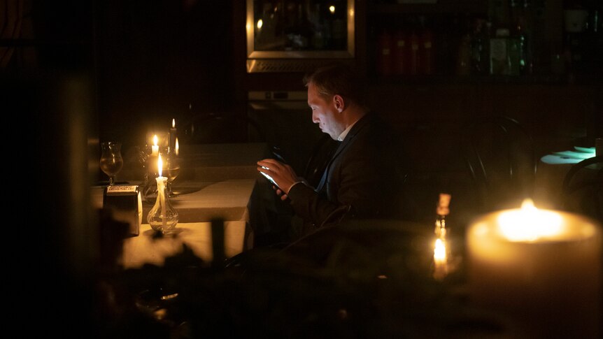 A man sits in a cafe and is illumted by candels and a smartphone screen during a blackout.