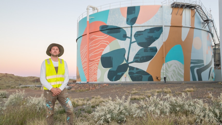 A man in high vis and a hat stands in front of a colourful abstract mural on a water tank