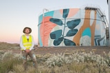 A man in high vis and a hat stands in front of a colourful abstract mural on a water tank