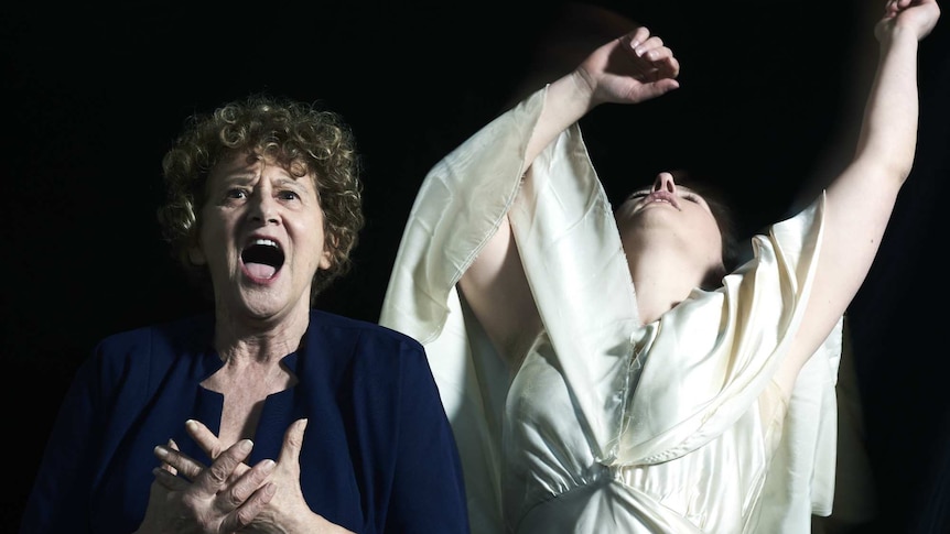 Two women in front of a black backdrop. The woman on the left sings as the woman on the right leans back with arms raised.