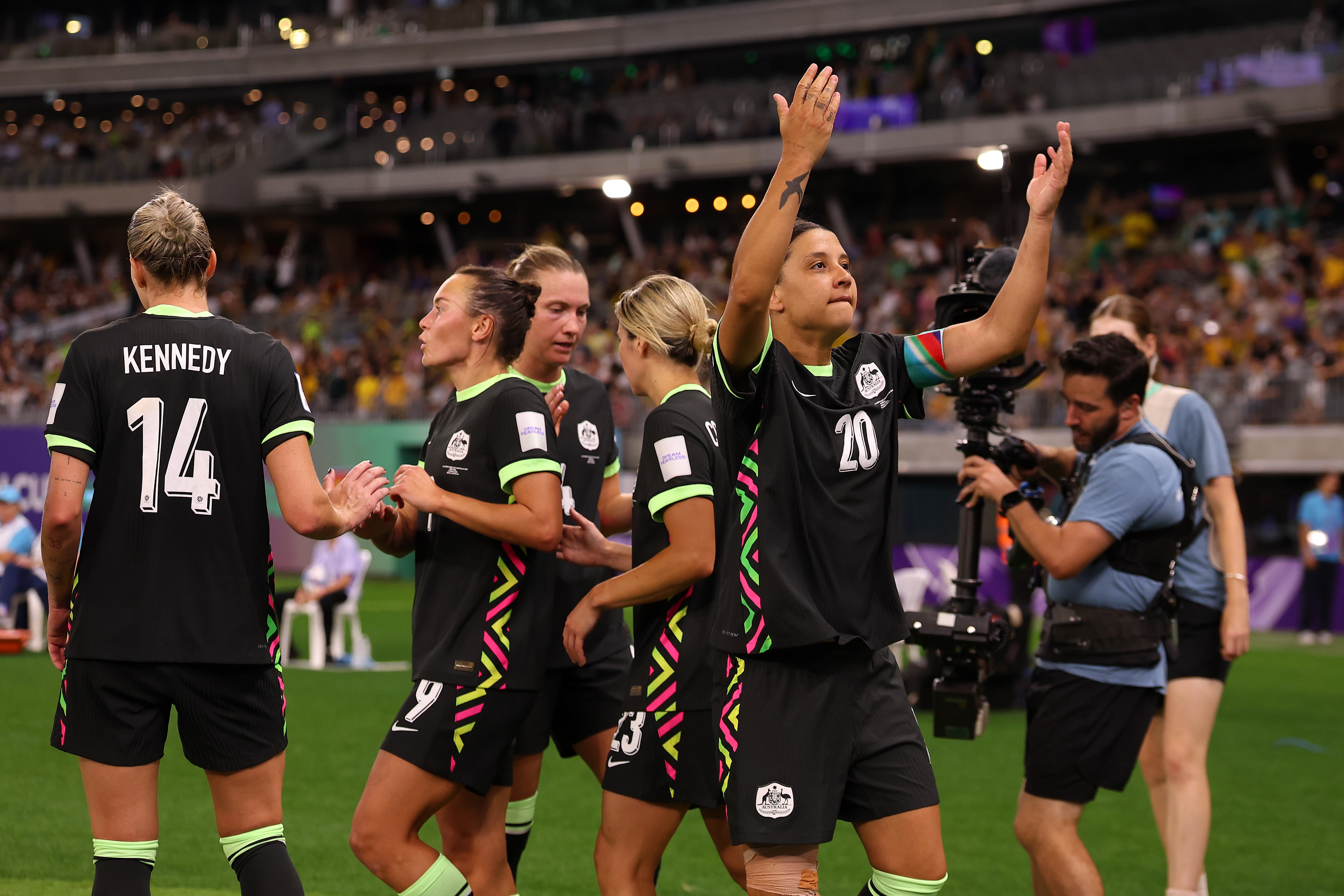 A soccer player in green holds up her hands in the air with teammates around her