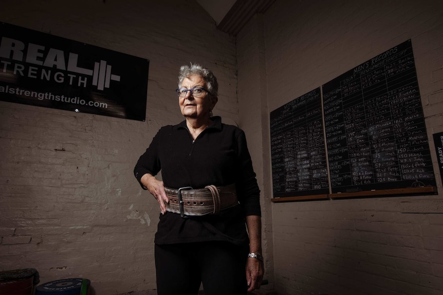 A woman in black with a weight belt on stands in front of a chalk board with her hand on her hip