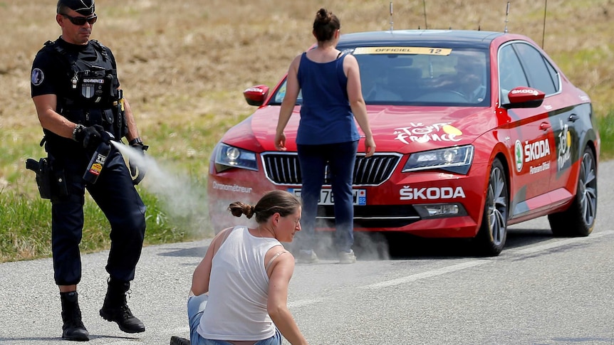 A police officer sprays a protester as another protester stands in front of a red car.
