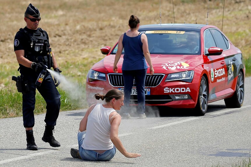 A police officer sprays a protester as another protester stands in front of a red car.