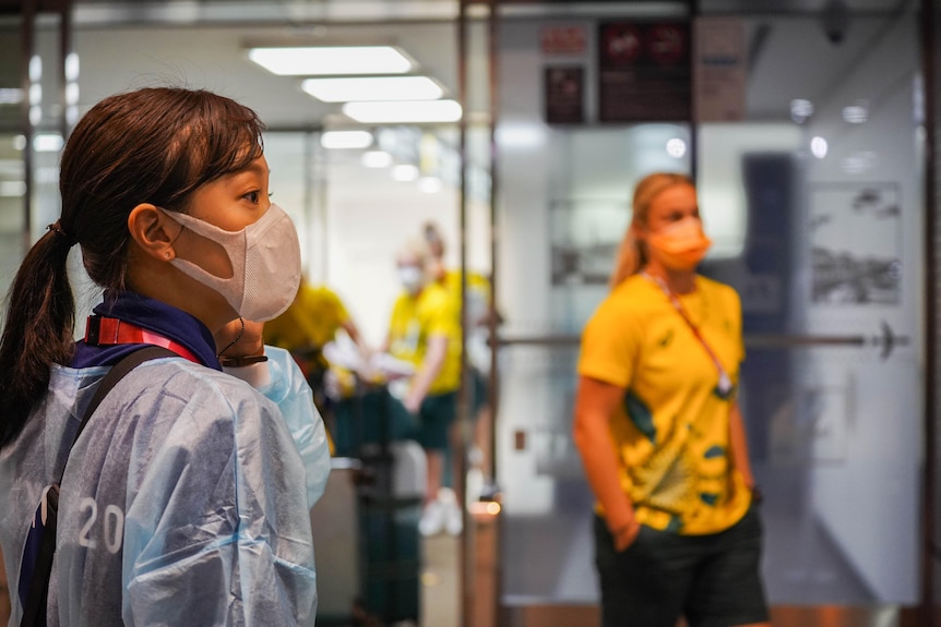 A masked woman in a Tokyo 2020 shirt looks off as an Australian female athlete lingers in the background 