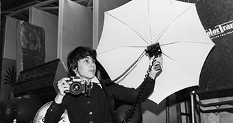 A woman stands and holds camera in right hand and flash umbrella in left.