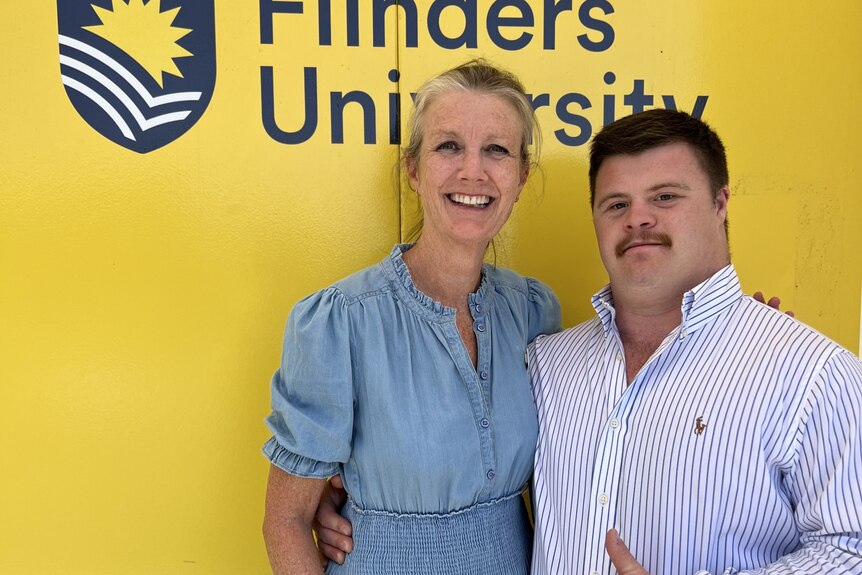 young man stands in front of a Flinders University banner with a woman wearing a blue dress