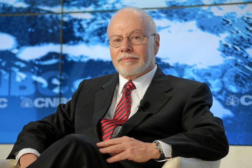 A man with bald head, white hair and a beard, siting in a chair, wearing a black suit, red tie and glasses.