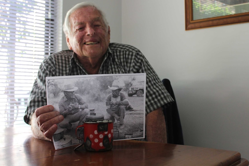 A white-haired man sitting at a table holds a photo.