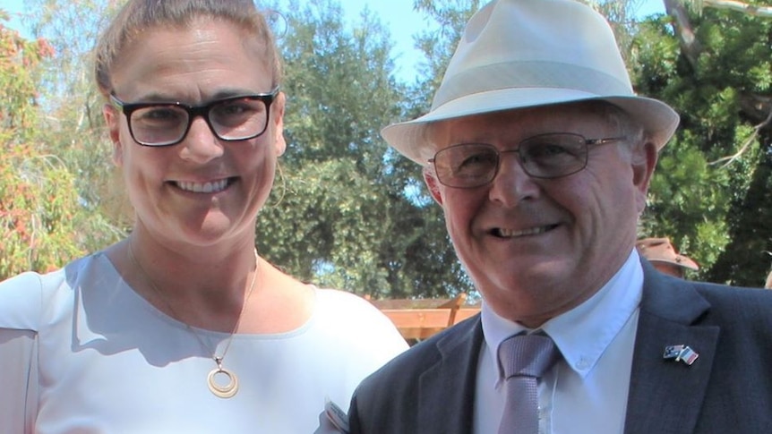 A middle-aged woman in glasses smiles while standing next to an older man wearing a hat and suit.