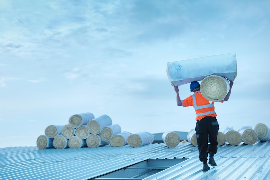 A man in high-vis carries a bale of insulation on a roof