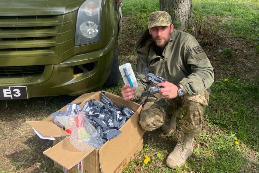 A man wearing a soldier's uniform squatting next to a box and a green van