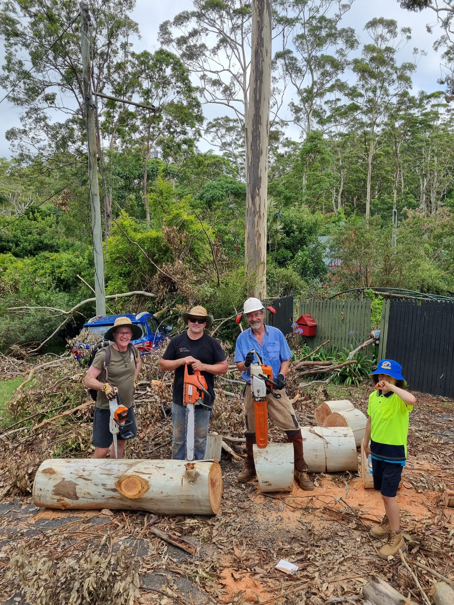 three men with chainsaws standing on top of a tree that has been cut up. A child stands next to them