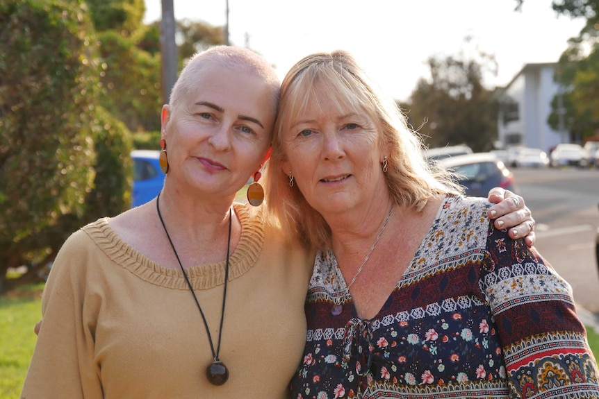 Two women in their 50s standing on a street looking at the camera with their arms around each other.