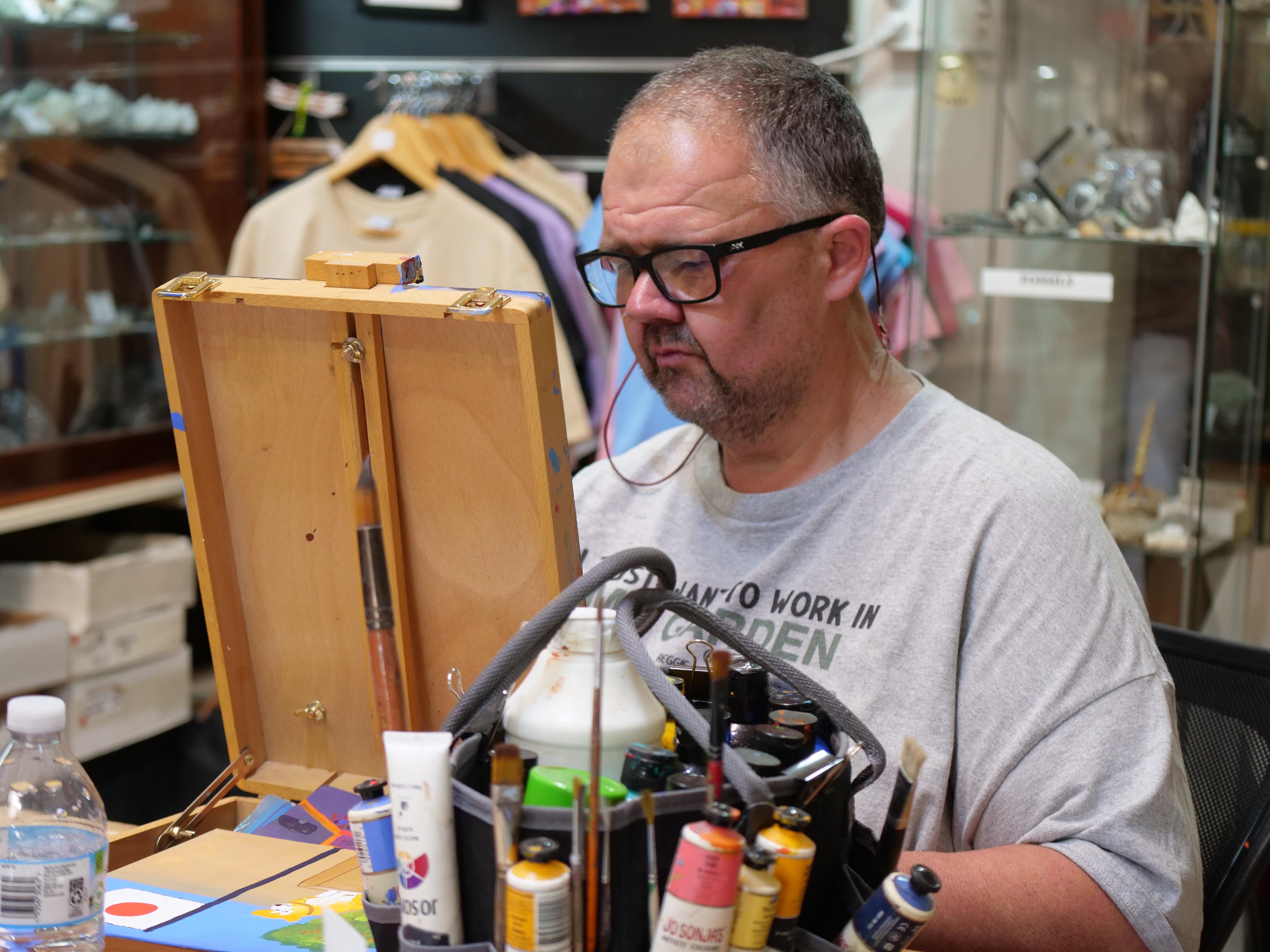 Un hombre con camisa gris y gafas negras mira un pequeño caballete. 