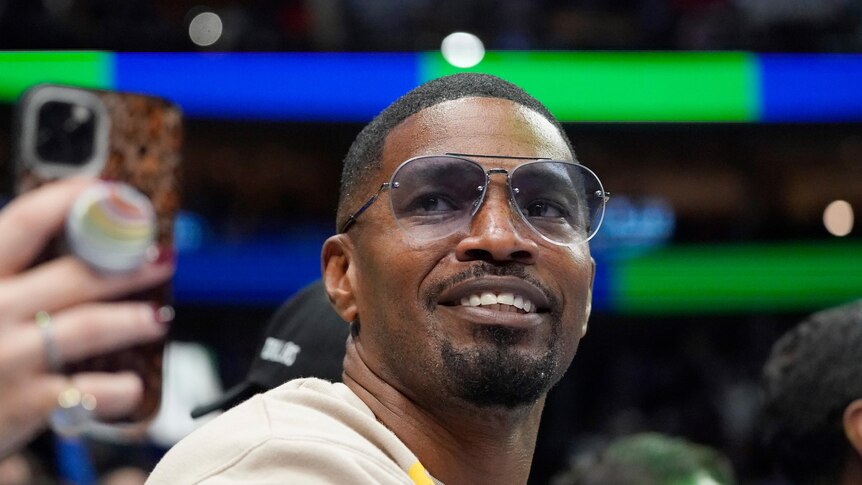 Jamie Foxx smiles while wearing sunglasses courtside at a basketball game