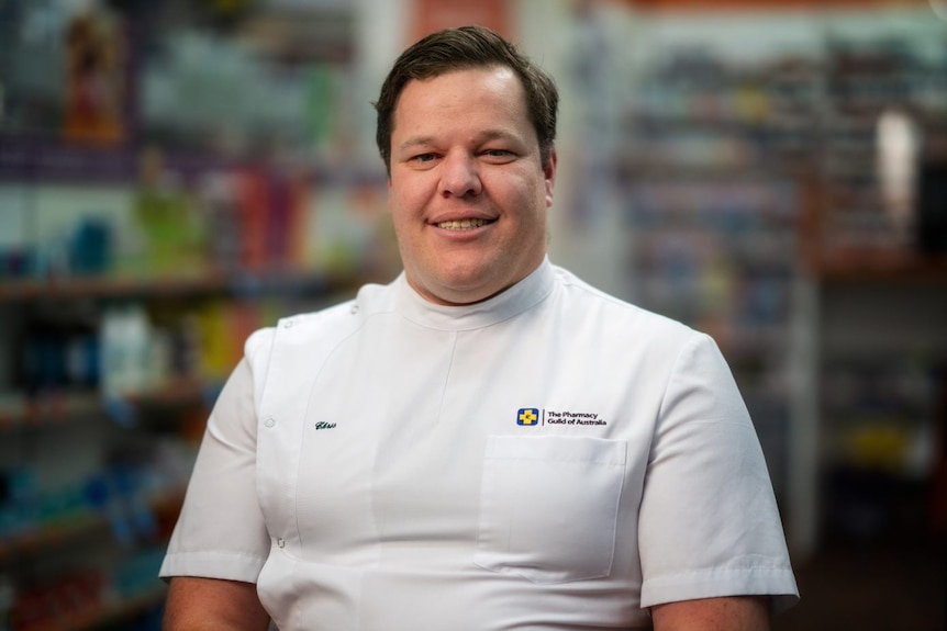 Man in a white lab shirt smiling in a pharmacy.