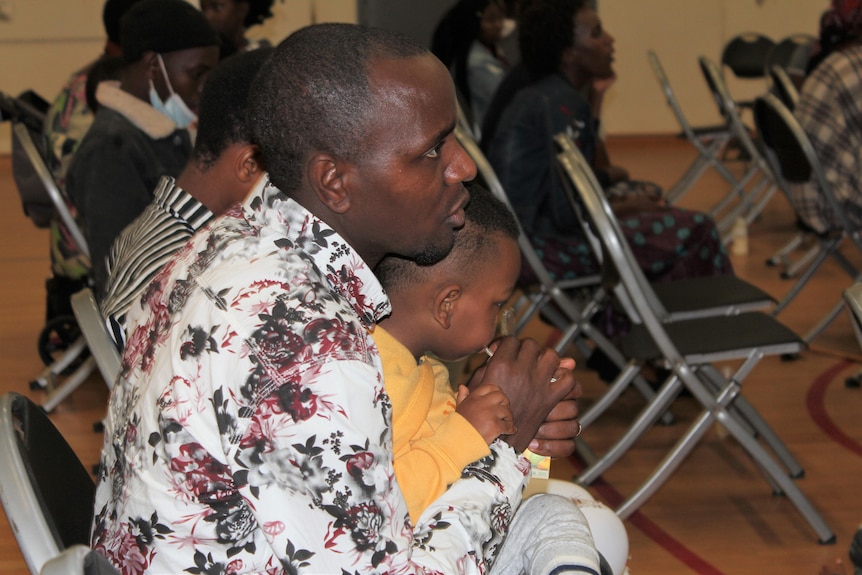 A man sits in a chair with a child in his lap and look ahead at a choir that's out of shot. 