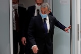 A man in suit wears a face mask as he pushes a door open to leave a room, with two other men following.