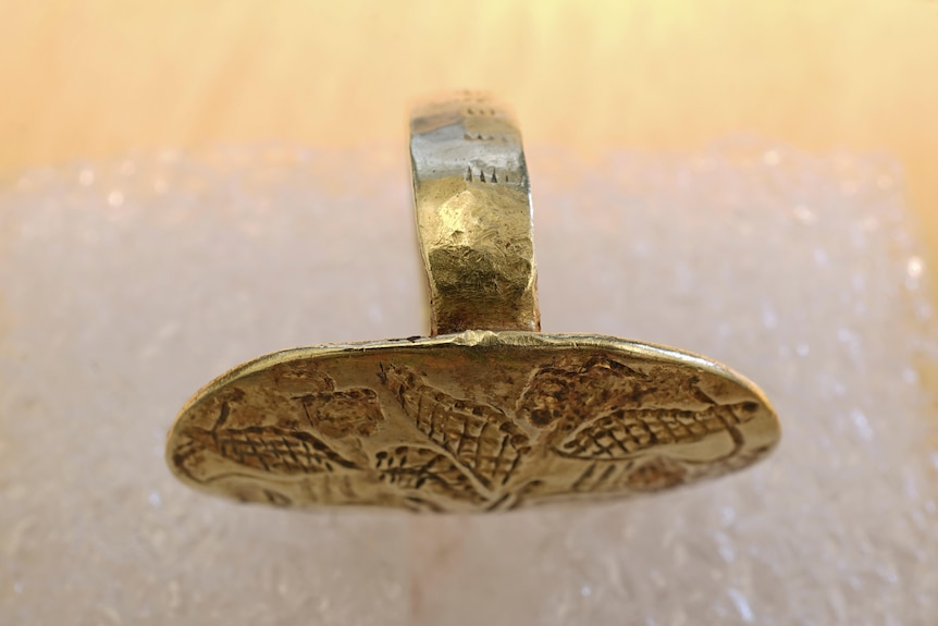 Oval-shape gold ring engraved with two facing sphinxes set on a white background.