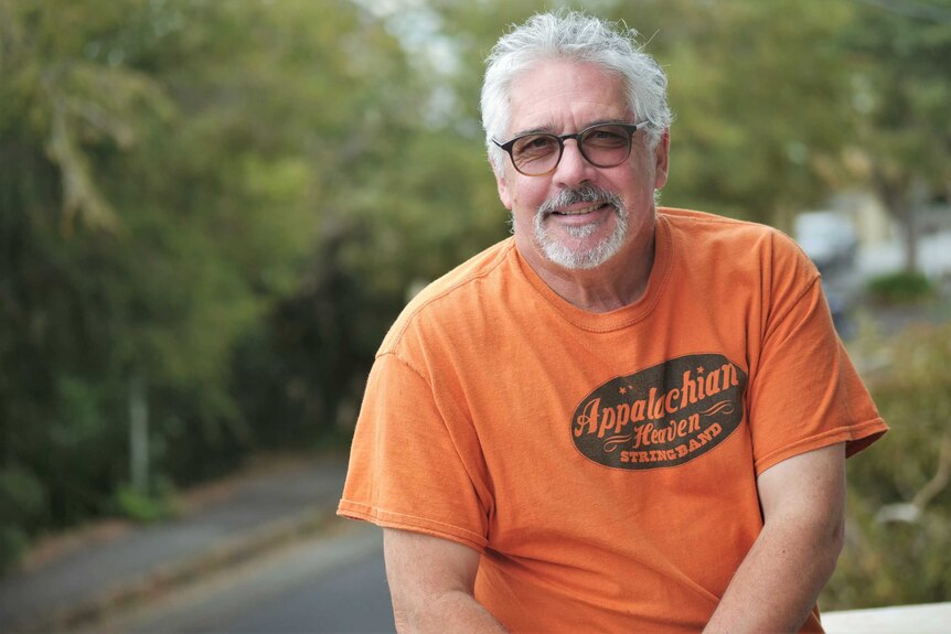 A bespectacled man with grey hair and a goatee wearing an orange shirt.