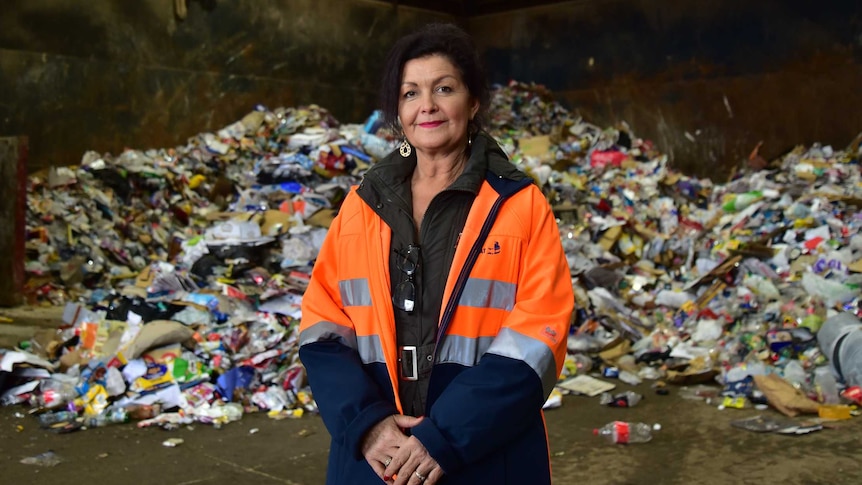 A woman in a high-vis jacket pictured standing in front of a large pile of rubbish in a warehouse.