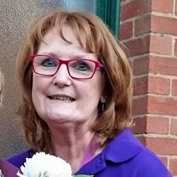 two older women wearing spectacles, one on left with short blonde hair and one on right with red bob holding flowers