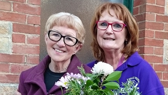 two older women wearing spectacles, one on left with short blonde hair and one on right with red bob holding flowers