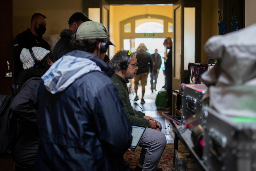 A man wearing headphones watches a screen as people stand around, also watching.