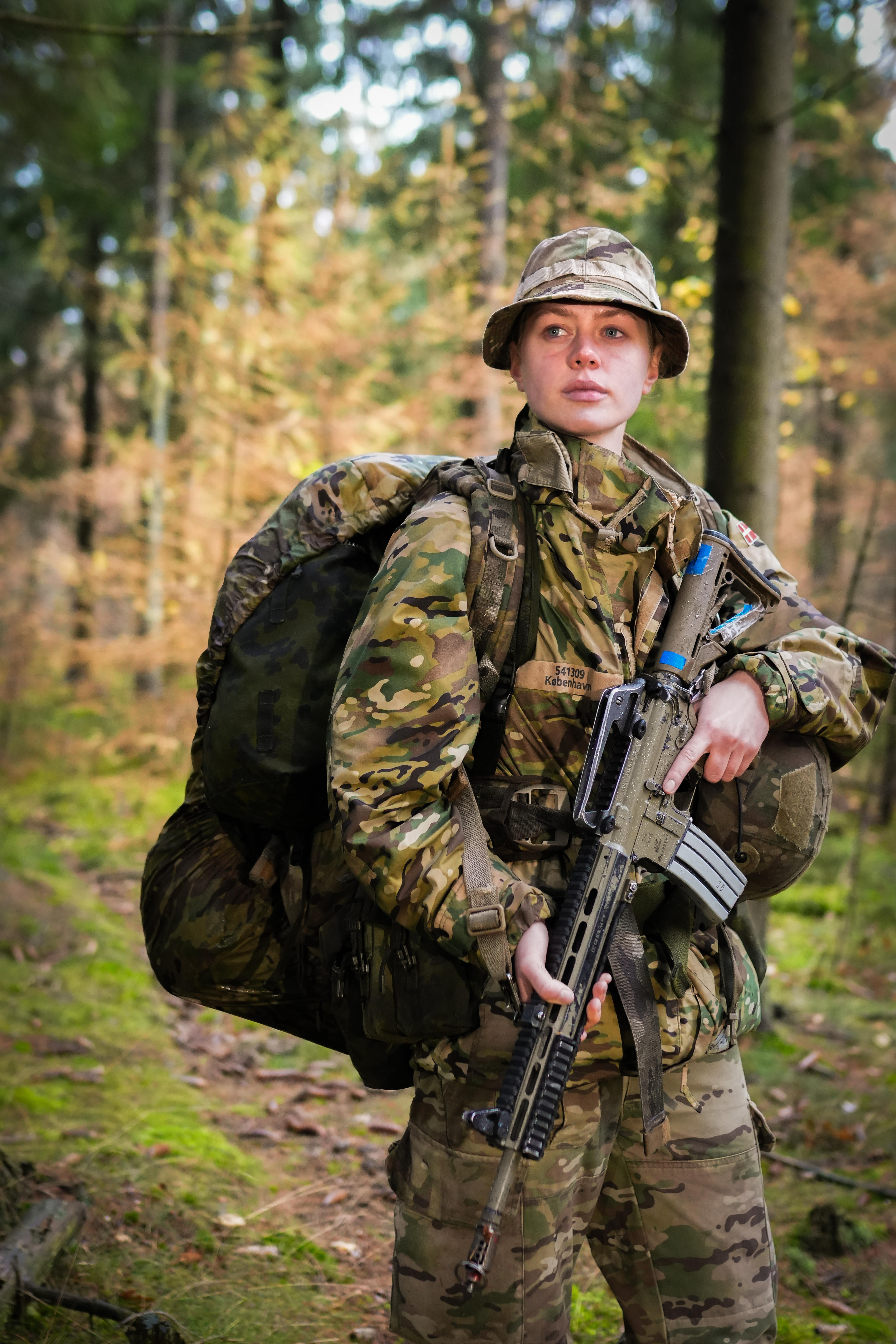 A young woman in khaki camouflage, holding a rifle in the woods.