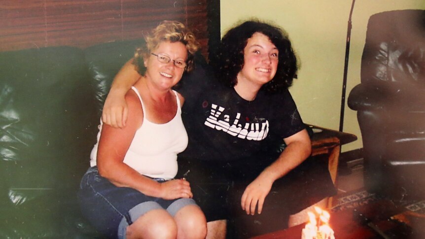 A woman and her son sitting next to each other on a couch, his arm around her shoulders.