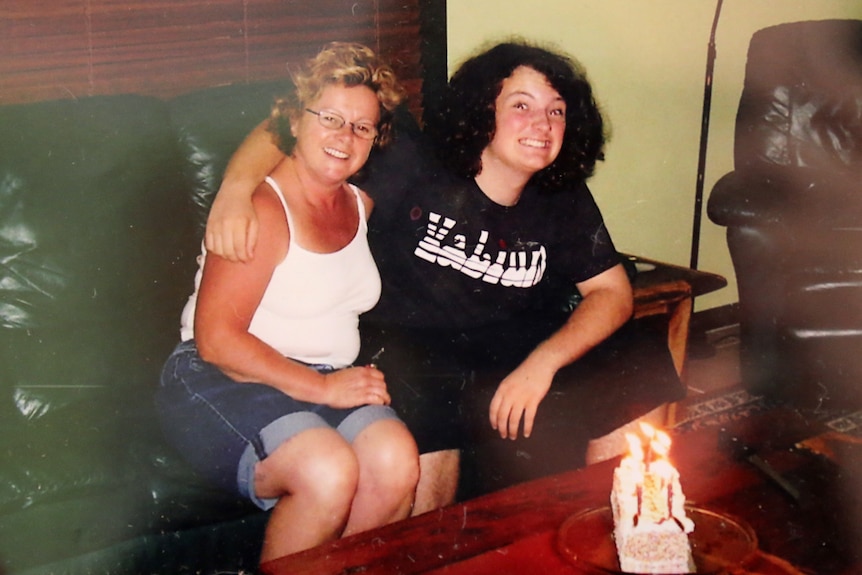 A woman and her son sitting next to each other on a couch, his arm around her shoulders.